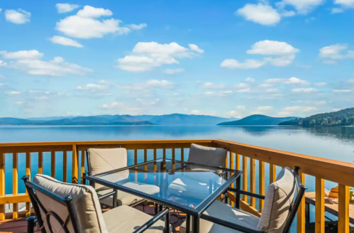 A serene lakeside view from a deck with a glass table and chairs, surrounded by mountains and a blue sky.